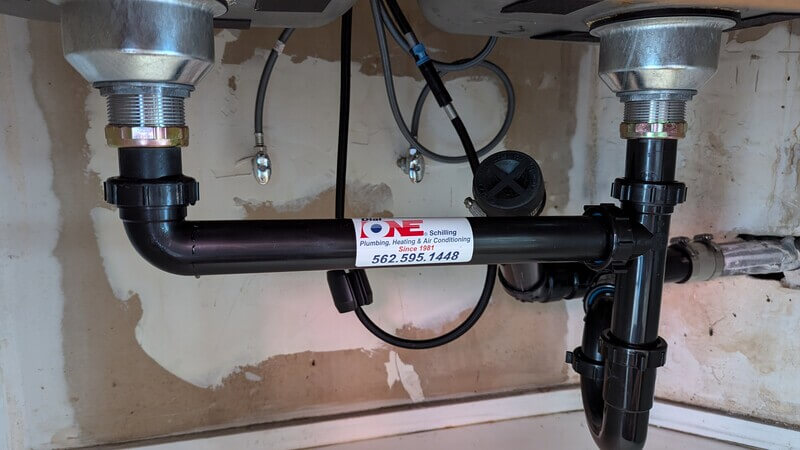 Under-sink drain installation with black ABS piping labeled Dial ONE Schilling, showing dual-basin connections and clean setup.