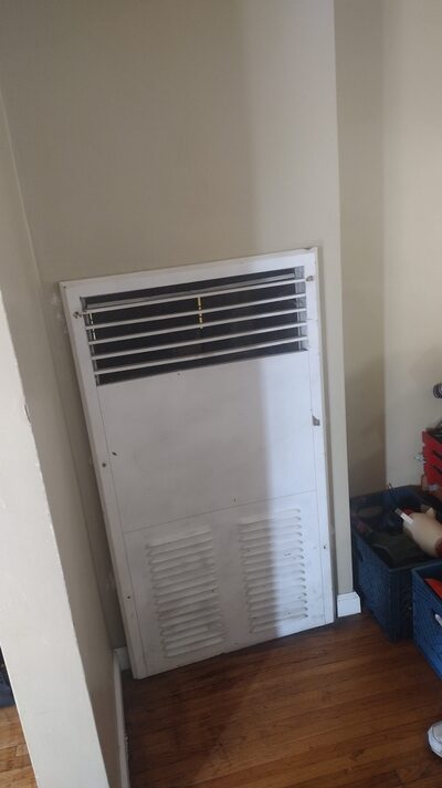 Wall-mounted gas heater with upper vent grille, installed in hallway of older home.