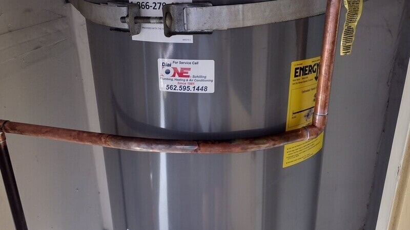 Residential water heater installation with copper piping, labeled with Dial ONE Schilling service sticker and energy efficiency guide.