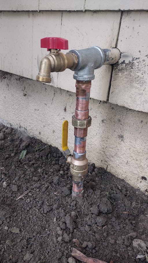 New hose bib installation with red handle and vertical copper piping, featuring a ball shutoff valve at the base in exposed soil.