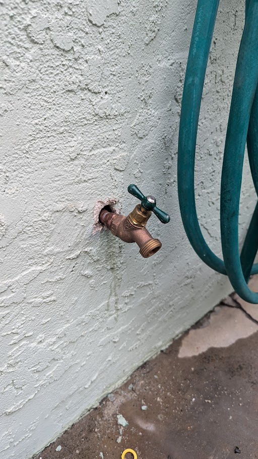 Wall-mounted hose bib with green handle, professionally installed into stucco exterior.