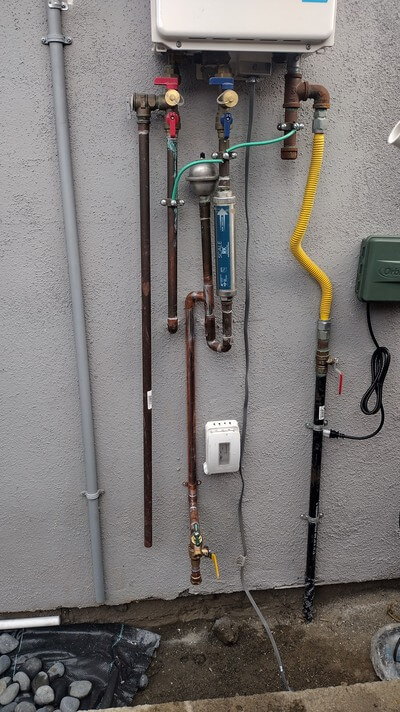 Exterior view of tankless water heater installation with gas, water, and filtration connections.