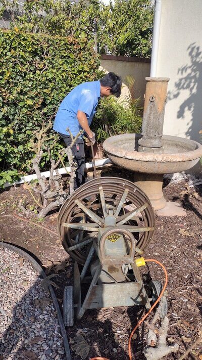 Dial ONE Schilling plumber using drain machine for outdoor sewer line near garden fountain.