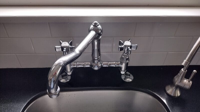 Close-up of vintage-style kitchen faucet with cross handles and polished chrome finish, installed on black countertop with white subway tile backsplash.