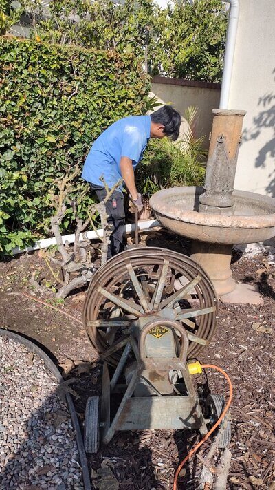 Plumber using drain snake machine near garden fountain.