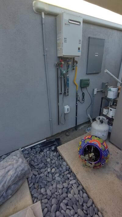 Outdoor tankless water heater with visible plumbing and gas lines against gray stucco wall.