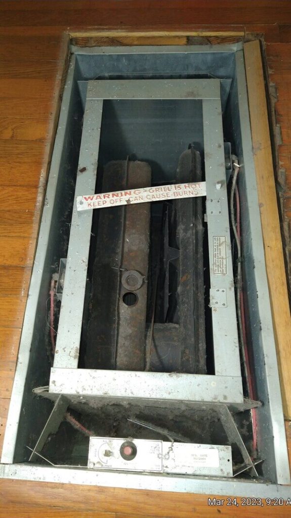 Open floor furnace showing internal burners and warning label reading “Grill is hot—keep off,” photographed during repair in a Lakewood, CA home by Dial ONE Schilling.