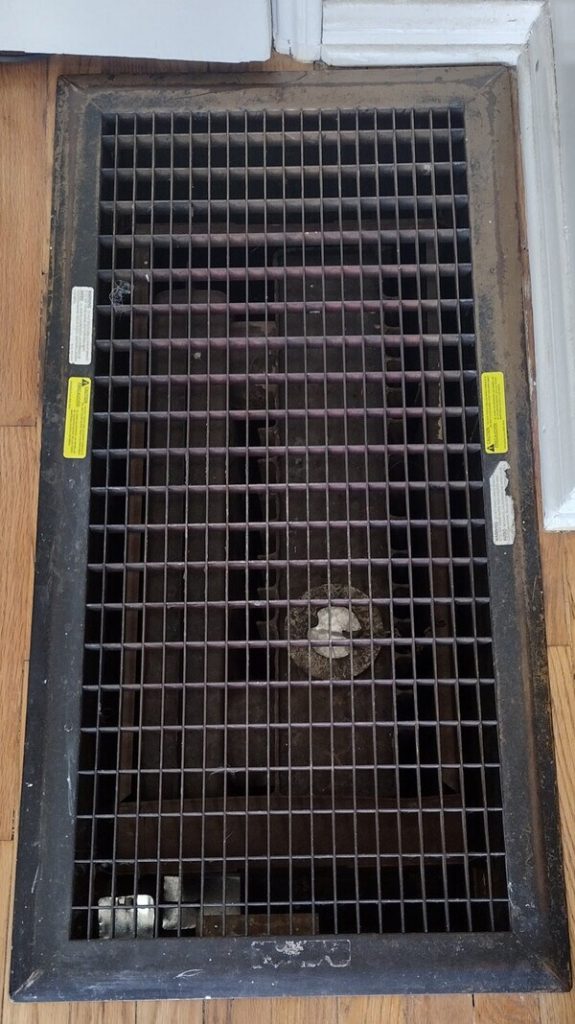 Floor furnace vent grille installed in older Lakewood home, showing metal grate and warning labels prior to service by Dial ONE Schilling.